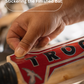 Hand applying a red and black 'TROGON' brand sticker to a wooden cricket bat.