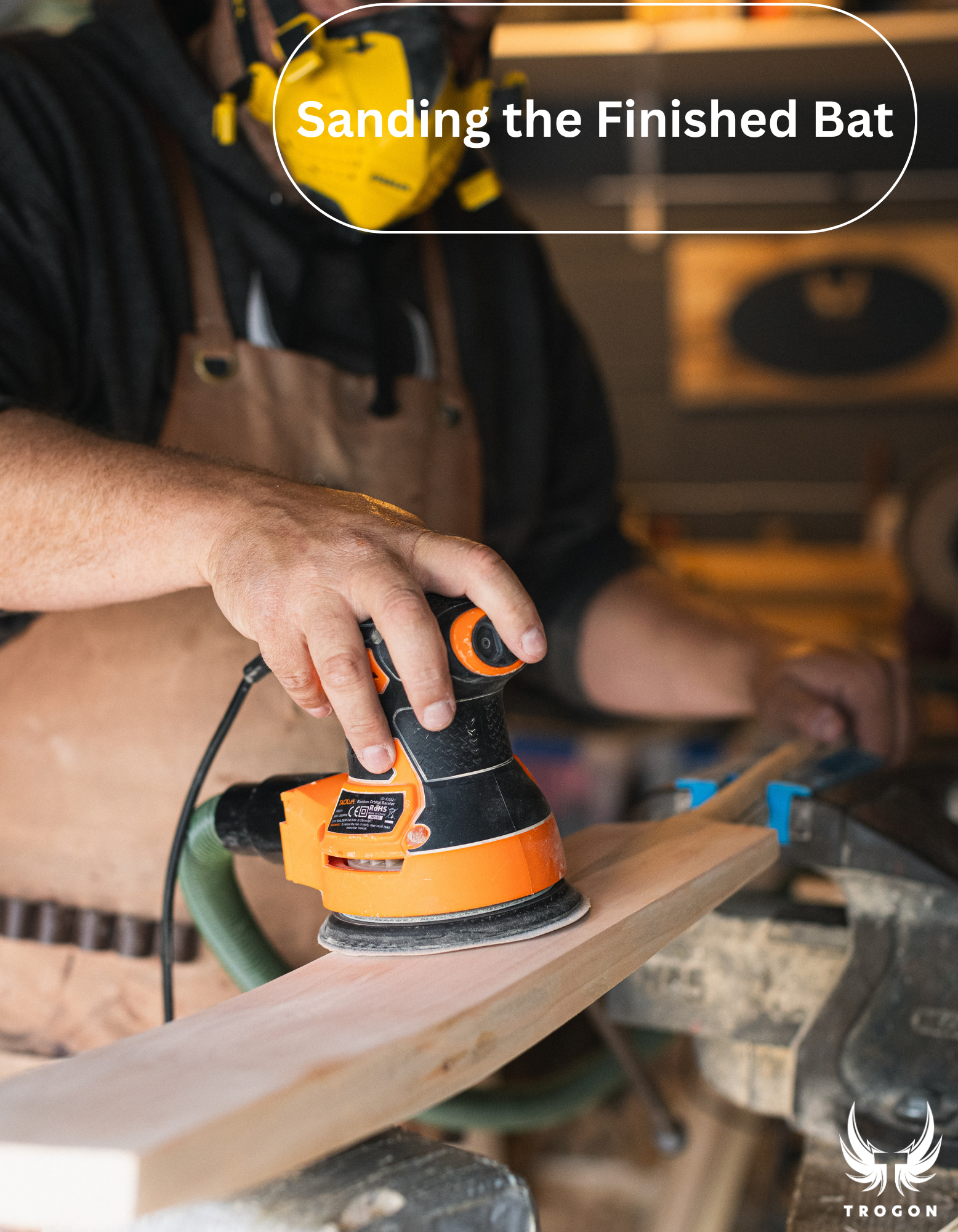 Person sanding a wooden Trogon cricket bat with a sander, wearing a yellow helmet and apron.