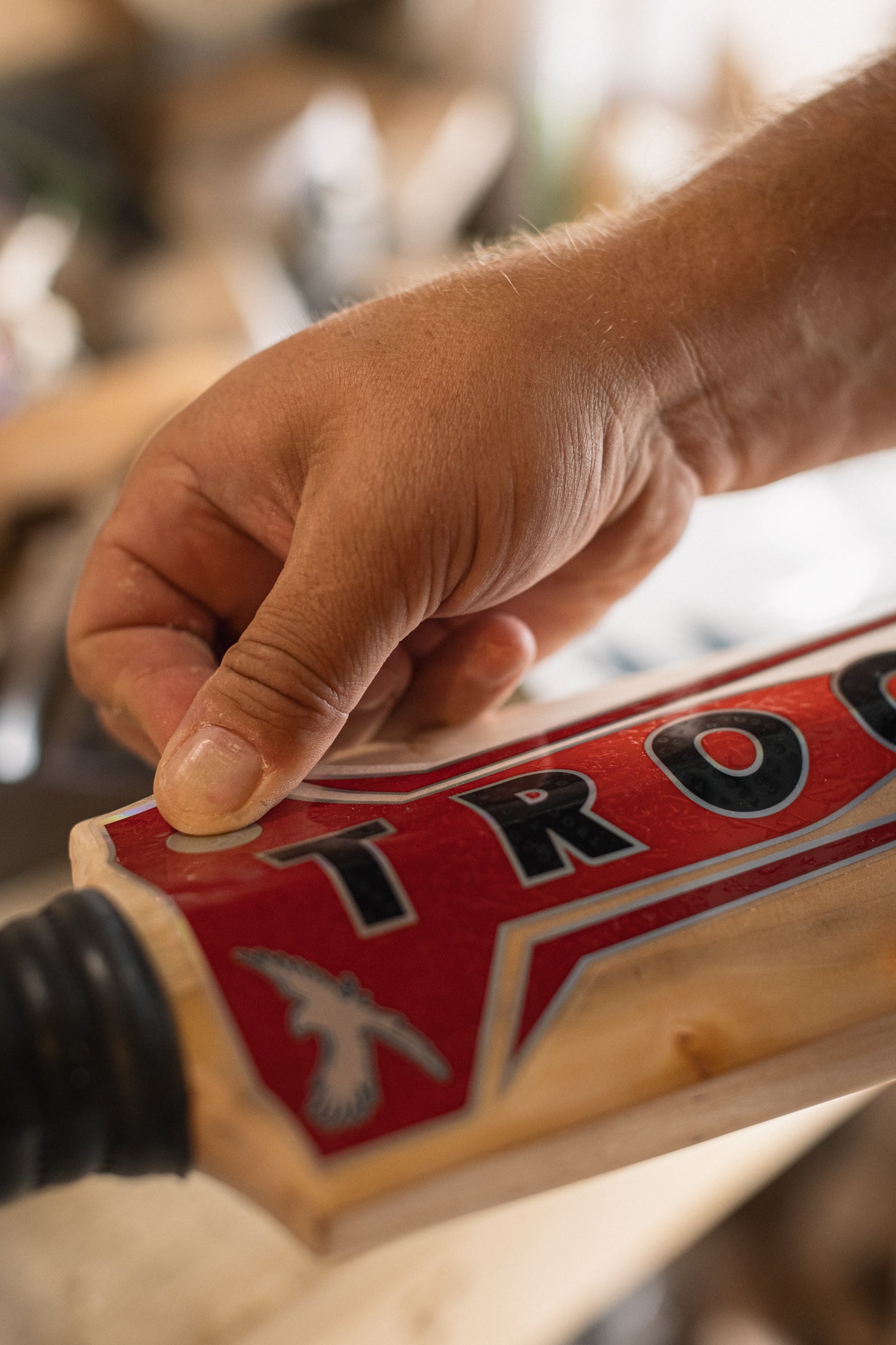 Hand applying a 'Trogon Cricket' brand label to a English willow cricket bat.